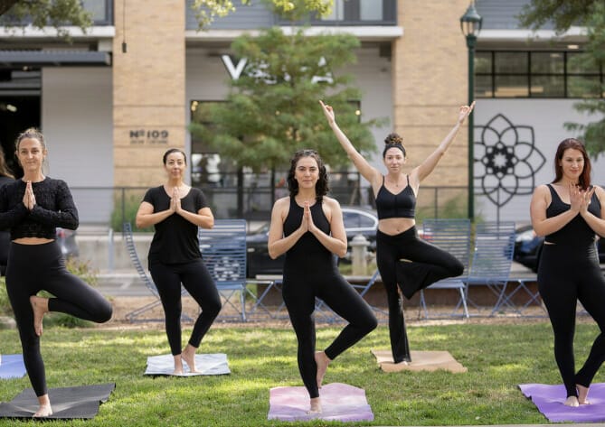 A group of yogi women doing a yoga class on the lawn of the MVP studio. There is a visible sign that says "yoga", and the MVP mandala logo on the wall of the white building.