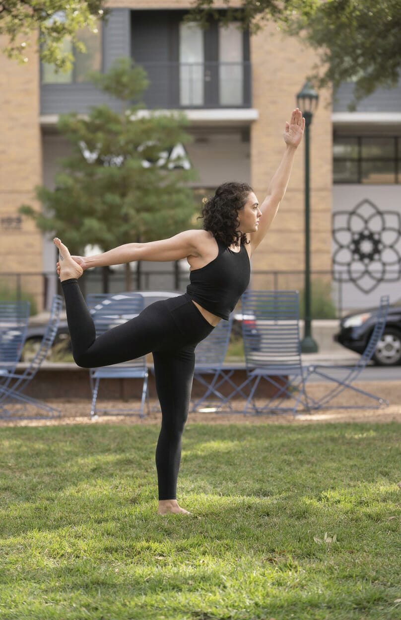 A woman yogi doing yoga on the lawn of the MVP studio. She is balanced on her left leg, and holding her right leg up and behind her.