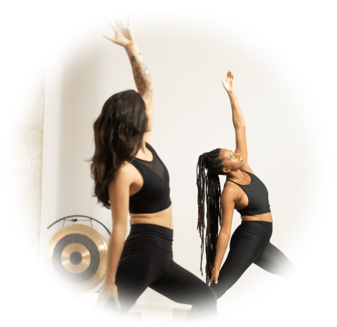 Two women yogis are in a yoga class. Their right arms are held straight up in the air.